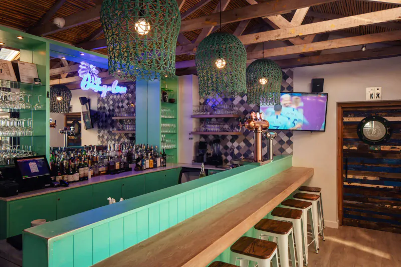 The Bungalow bar with neon sign, green pendant lights, wooden ceiling, stocked shelves, stools, and TV in Plettenberg Bay.