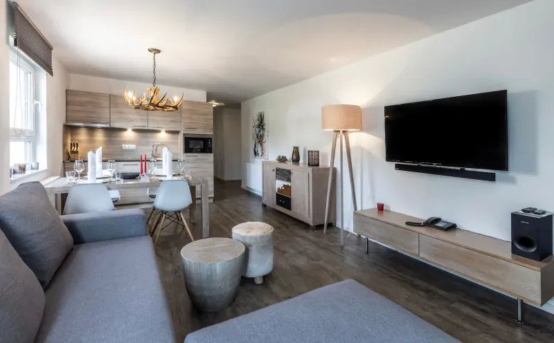 Modern open-plan living room with gray sofa, wooden stools, TV, floor lamp, and adjacent light wood kitchen.