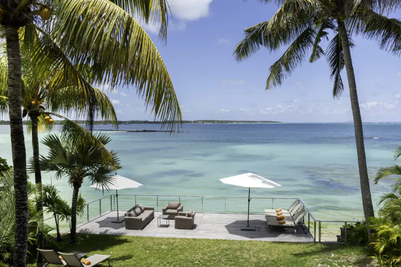Luxury villa deck with loungers and white umbrellas overlooking turquoise Mauritius beach and palm trees