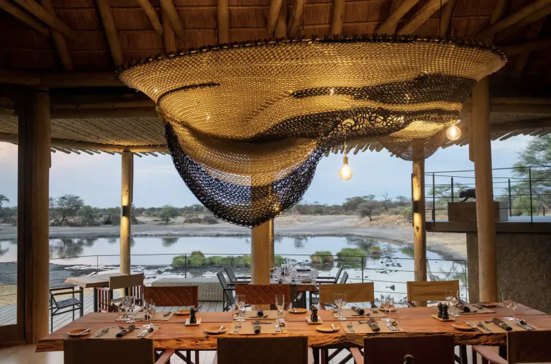 Luxury lodge dining room with woven chandelier, set table, and Etosha lake view through windows at Camp Kala.