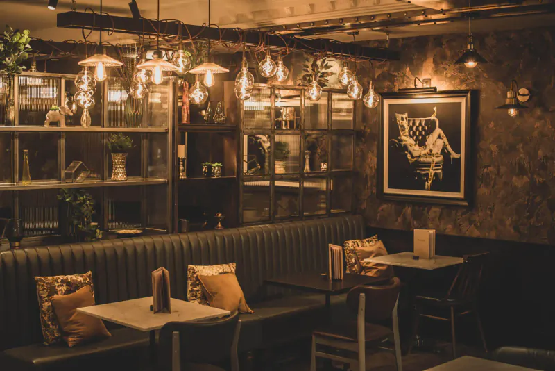 Cozy Six by Nico restaurant interior with warm pendant lights, metal shelves, abstract art, plush bench, and tables.