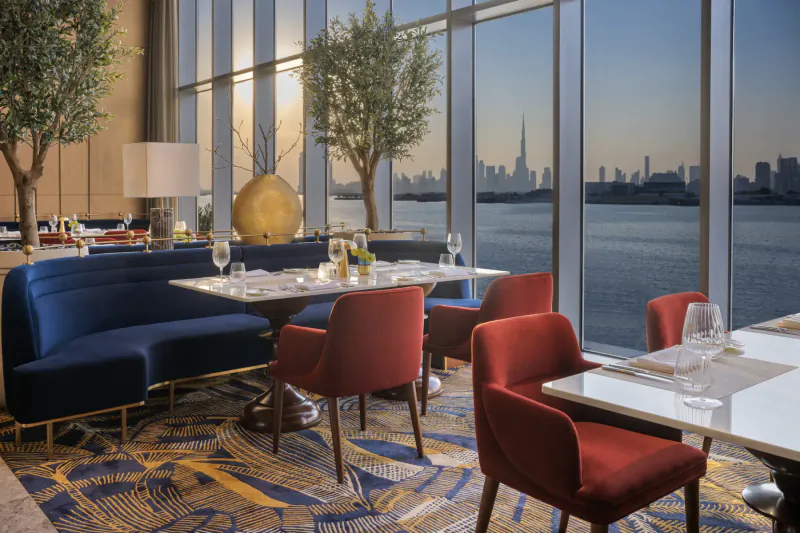 Elegant restaurant interior at dusk with blue velvet sofas, red chairs, white tables set with wine glasses, olive trees, and Burj Khalifa skyline view over creek.