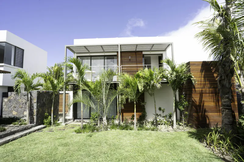 Modern tropical villa with white and wood facade, large glass windows, surrounded by palm trees and lush green lawn in Mauritius.