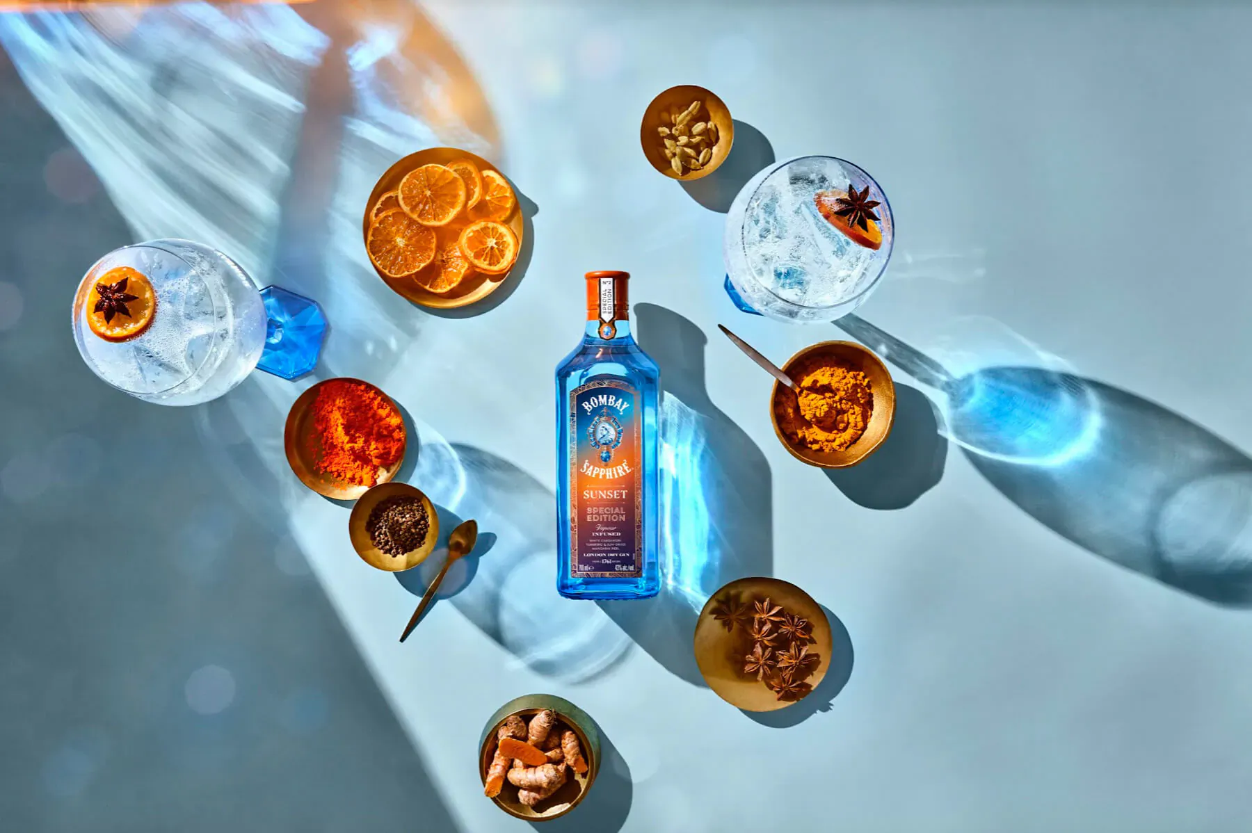 Bombay Sapphire gin bottle centered with gin & tonic, orange slices, star anise, and spices on blue surface