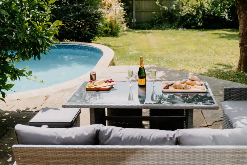 Outdoor garden table by pool with charcuterie board, champagne bottle, glasses, and cushions at Cotswolds country house.