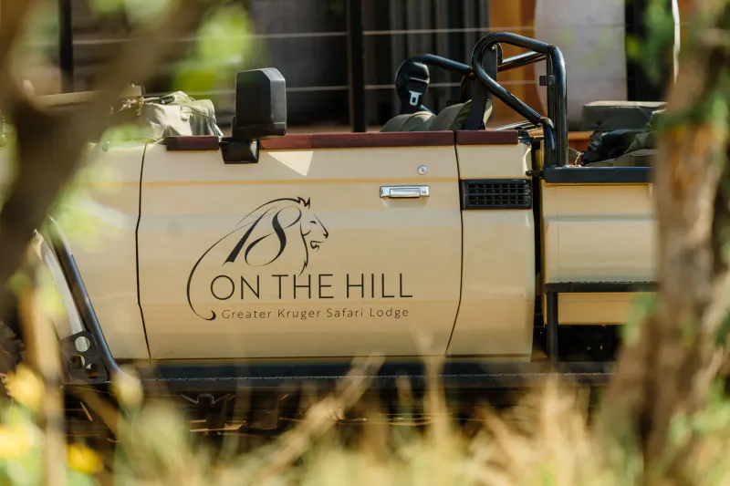 Beige Land Rover with 'On The Hill Greater Kruger Safari Lodge' logo on side, framed by green bush