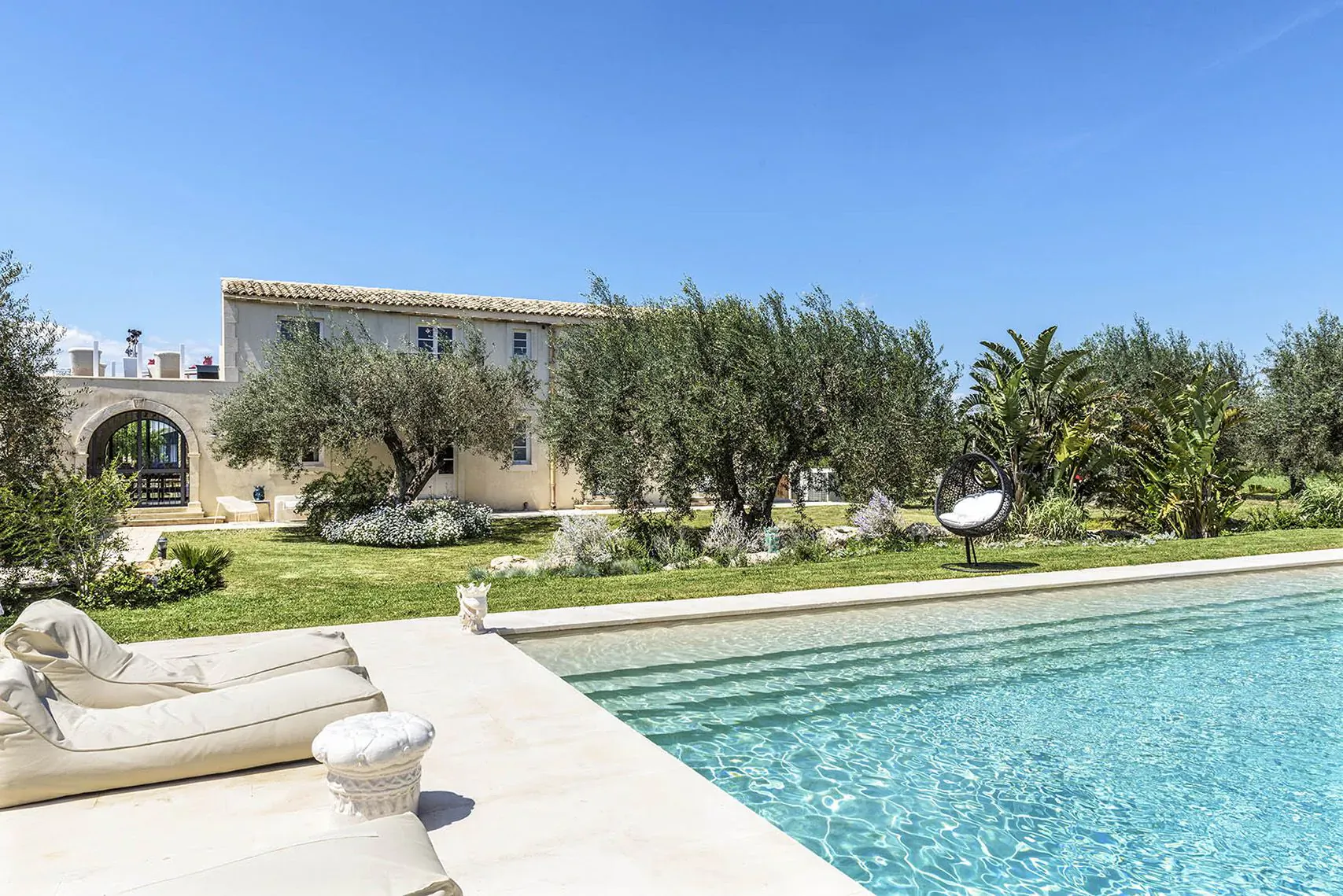 Zahir Country House in Sicily: beige stone villa, olive trees, palm, lush garden, infinity pool with loungers under blue sky
