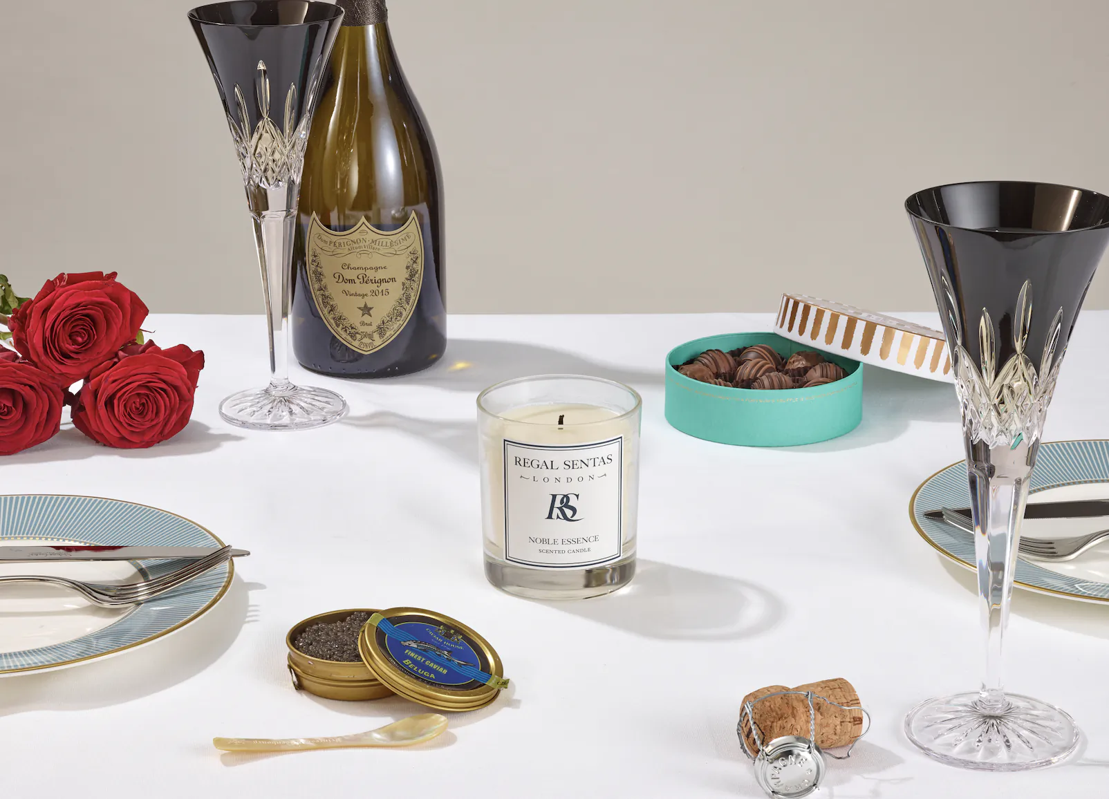 Elegant table setting with Dom Pérignon champagne, flutes, Diptyque '14' candle, caviar tin, chocolates, roses, and cork.
