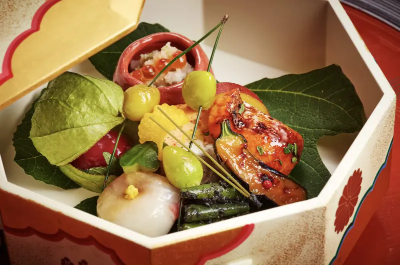Open lacquered bento box with kaiseki meal: grilled fish, asparagus, cherry tomatoes, physalis, and garnishes on leaves.