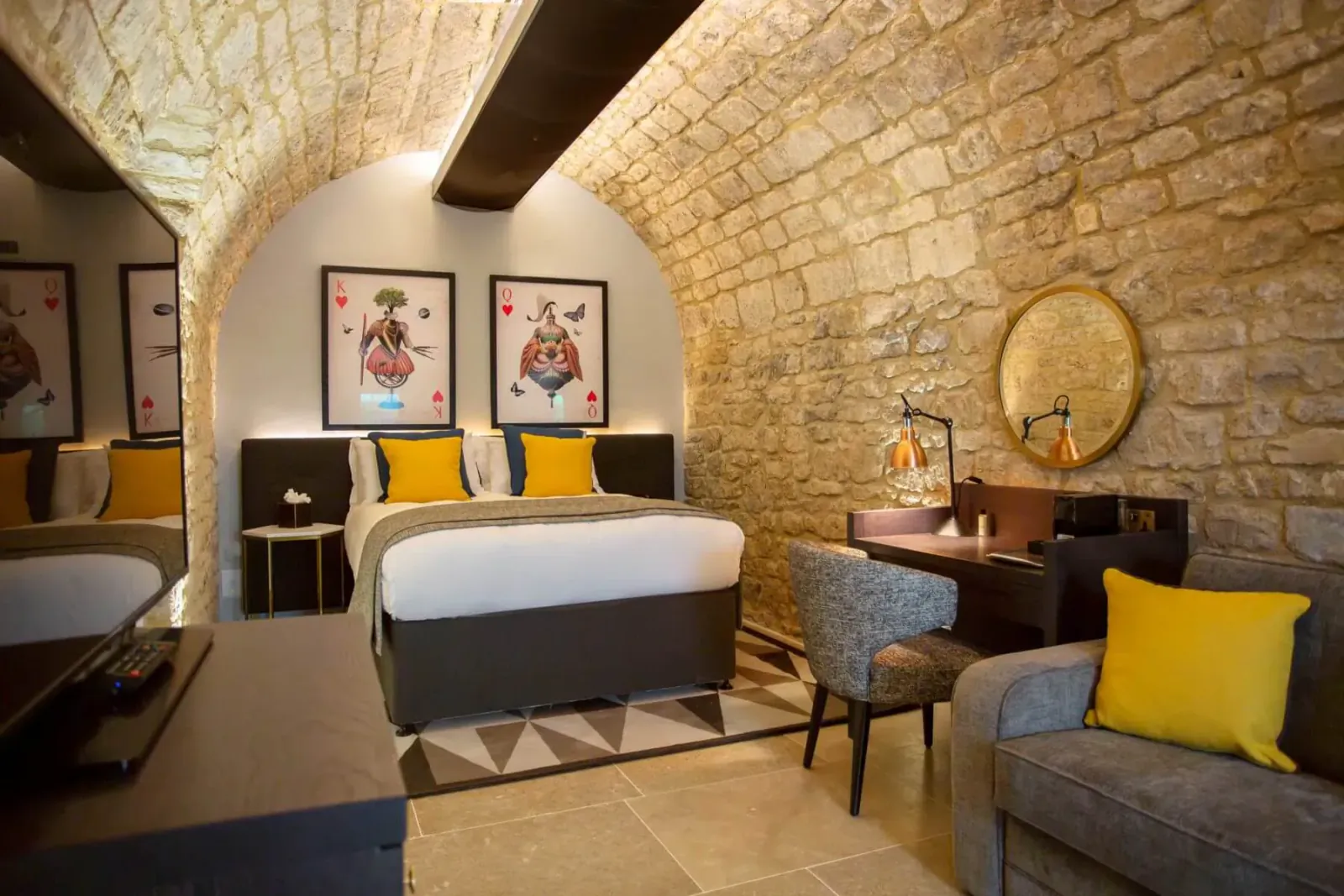Cozy underground vault hotel room with stone arches, gray bed, yellow pillows, desk, chair, and art.