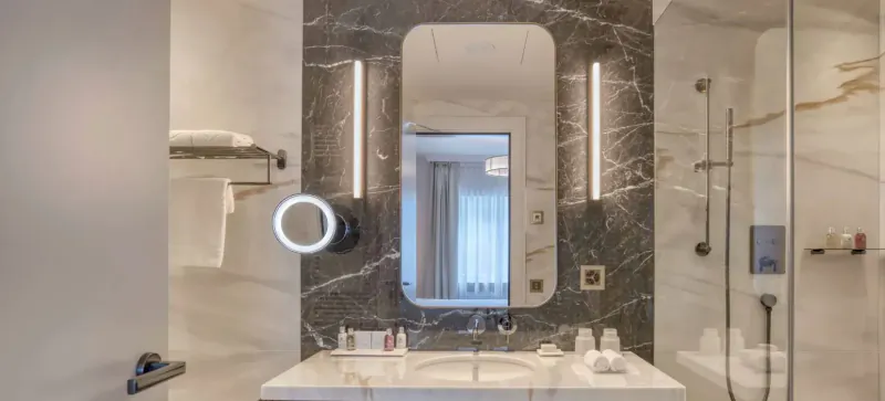Luxurious hotel bathroom vanity with marble walls, backlit mirror, towels, and glass shower at Grand Hotel Des Bains Kempinski St. Moritz