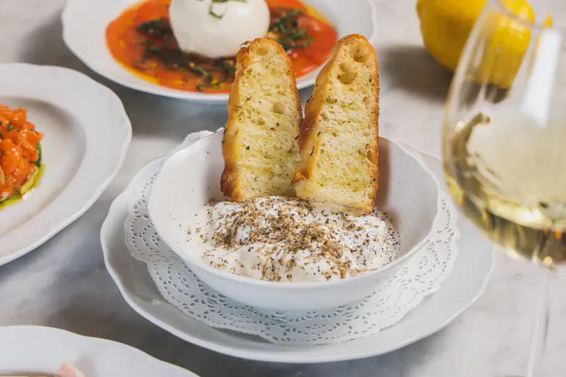 Two pieces of crusty bread in a bowl of creamy dip, with burrata salad, tomato sauce, and white wine glass nearby