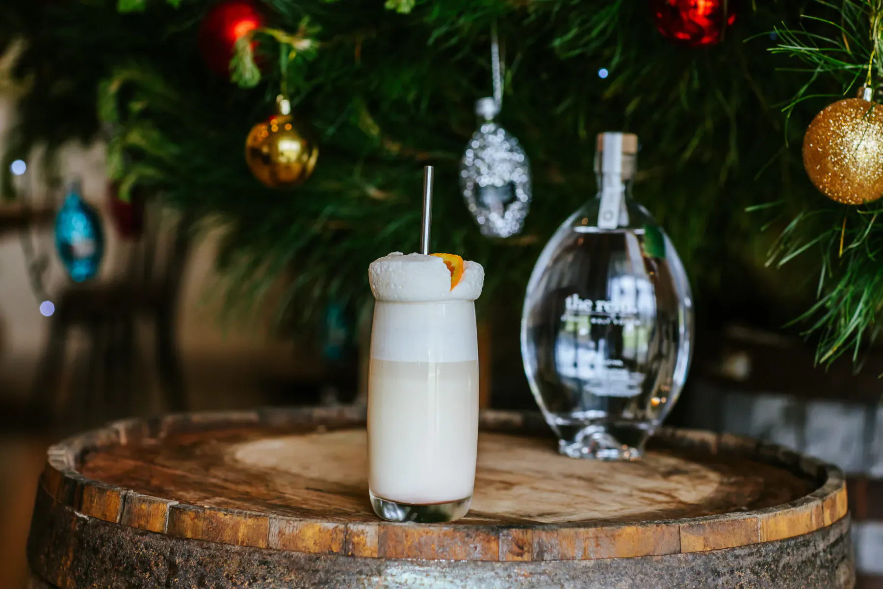 Creamy white Reid Single Malt Vodka cocktail topped with orange slice on barrel, beside clear bottle, with Christmas tree and ornaments.