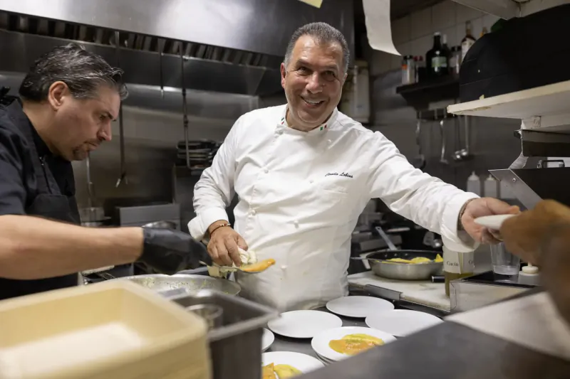 Two chefs in a professional kitchen collaborate, smiling chef in white uniform handing yellow food to black-clad chef amid plates and pans.