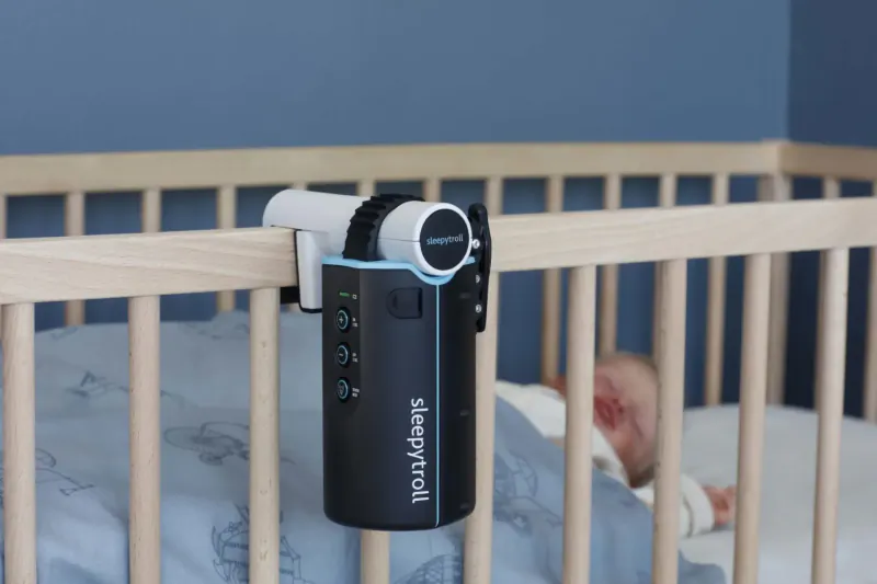 White Sensecam baby monitor with 'Snuza' branding clipped to wooden crib railing beside sleeping newborn.