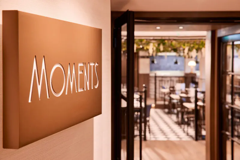 Moments sign on wall beside open doors to elegant restaurant with tables, chairs, and hanging plants on VIVA ENJOY ship.