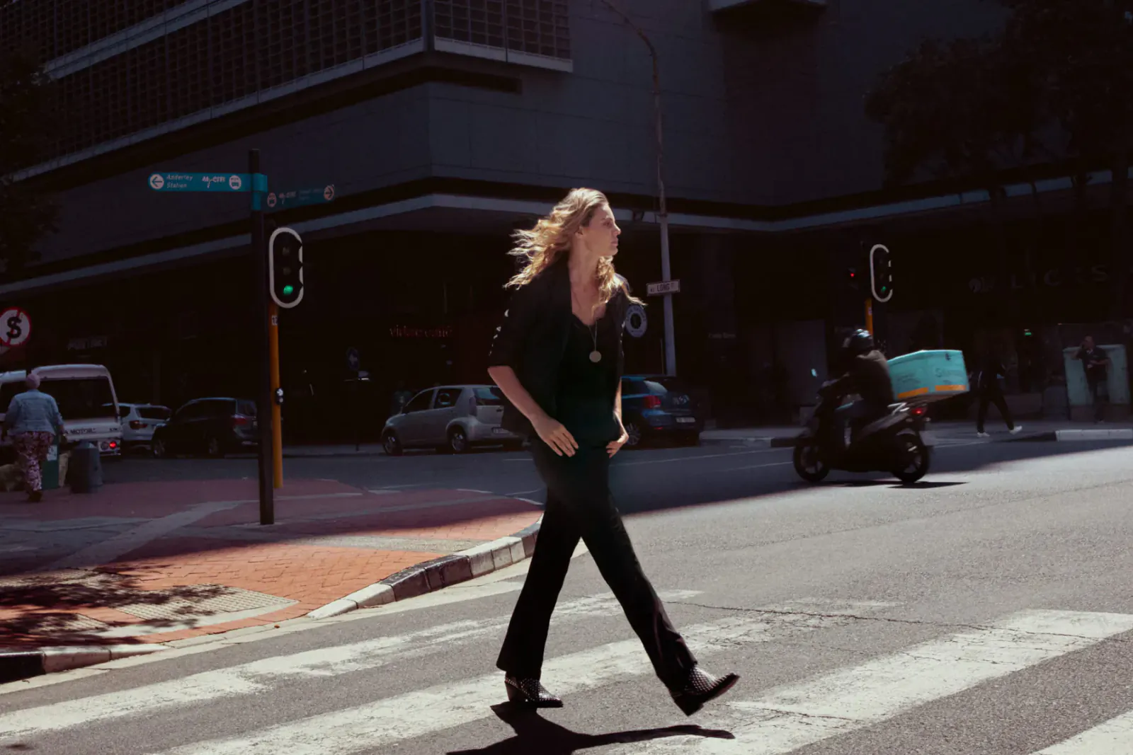 Blonde woman in black outfit with sophisticated urban style strides across city crosswalk at green light