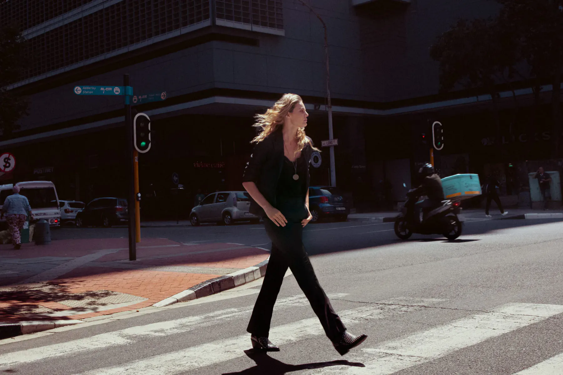 Blonde woman in black outfit with sophisticated urban style strides across city crosswalk at green light