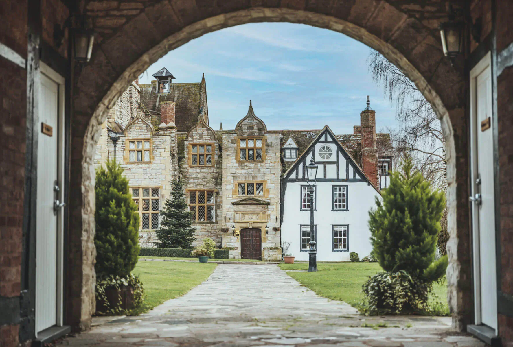 View through stone archway of Karma Salford Hall, Tudor-style mansion with half-timbered facade, gardens, and pathway in Vale of Evesham.
