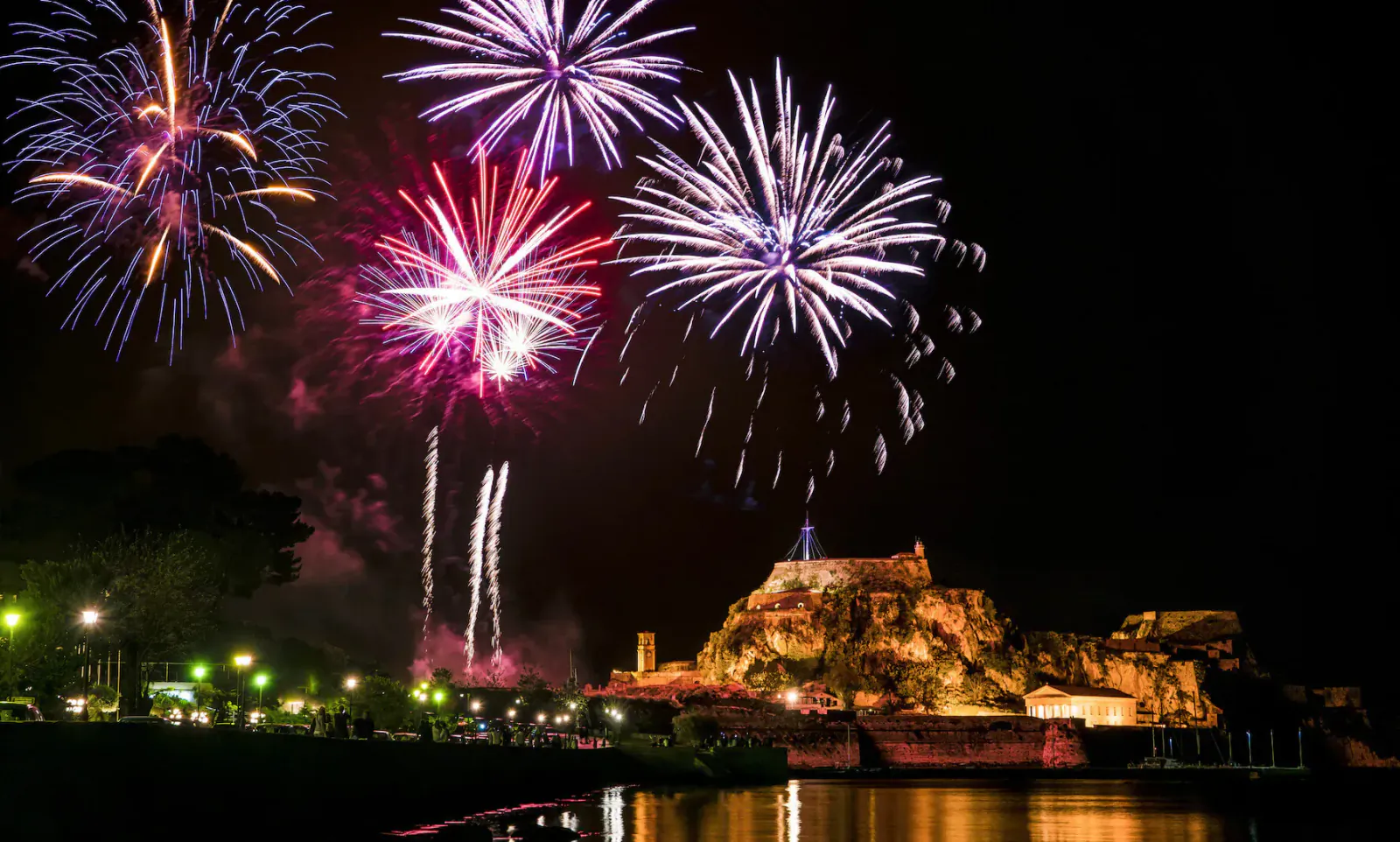 Vibrant red and purple fireworks exploding over a lit castle and waterfront during Easter holidays in Greece
