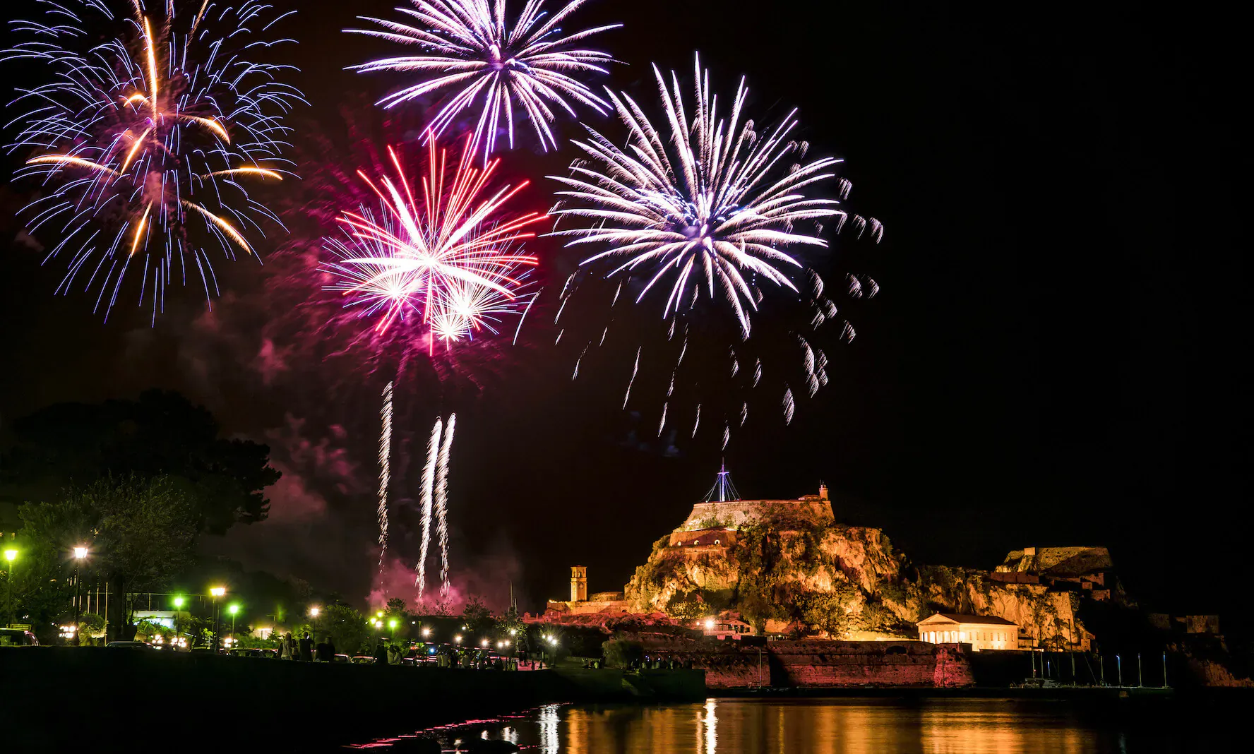 Vibrant red and purple fireworks exploding over a lit castle and waterfront during Easter holidays in Greece