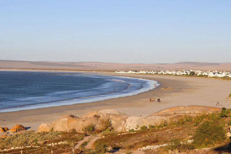 Aerial view of Paternoster beach with curving sandy shore, ocean waves, distant white houses, rocks, and tents under clear sky.
