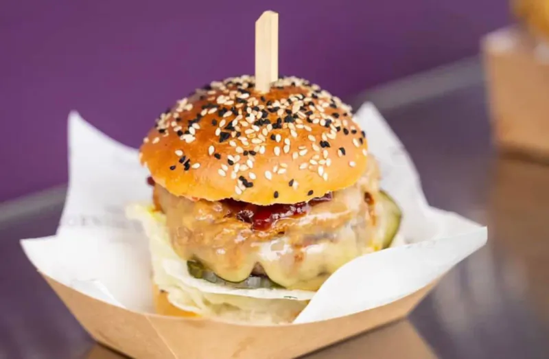Close-up of gourmet sesame seed burger with cheese, lettuce, pickles, and sauce on wooden skewer in paper tray