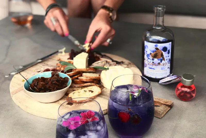 Woman slicing cheese on charcuterie board with Sugarbird Cape Fynbos Gin bottle and purple flower cocktails beside it.