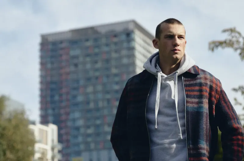 Man in white hoodie and plaid jacket stands confidently before tall Canary Wharf buildings on sunny day