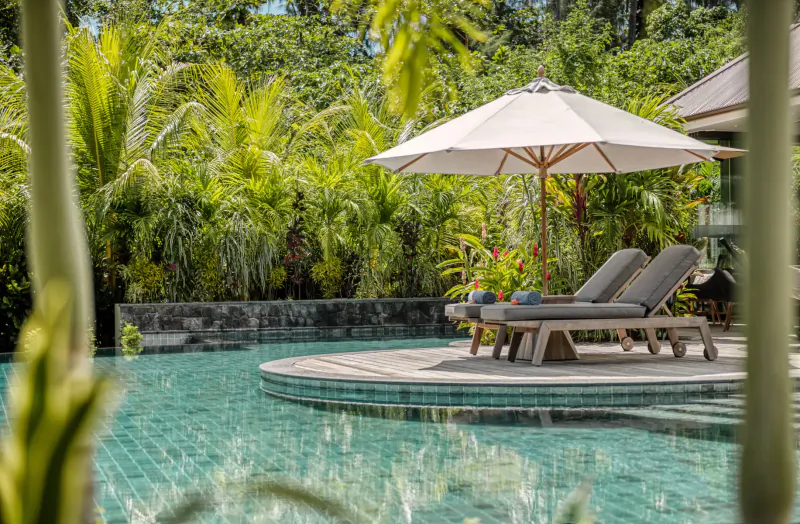 Two lounge chairs with pillows under a beige umbrella on a round pool deck at La Cigale Estate, surrounded by lush tropical greenery, Seychelles.