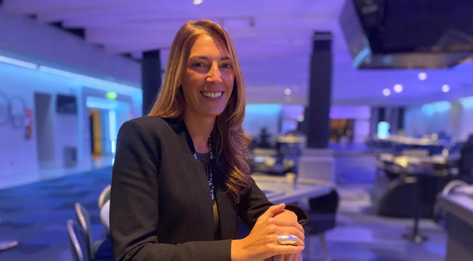 Paula Veiga Ortiz smiling in black blazer, seated at casino table with purple neon lighting, for Luxuria Lifestyle Spain feature.
