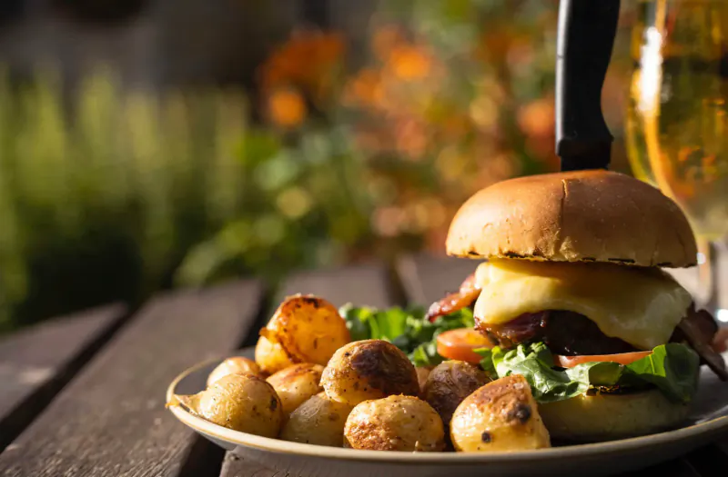 Bacon cheeseburger with lettuce, tomato, roasted potatoes on plate, beer glass, on wooden table in garden setting.