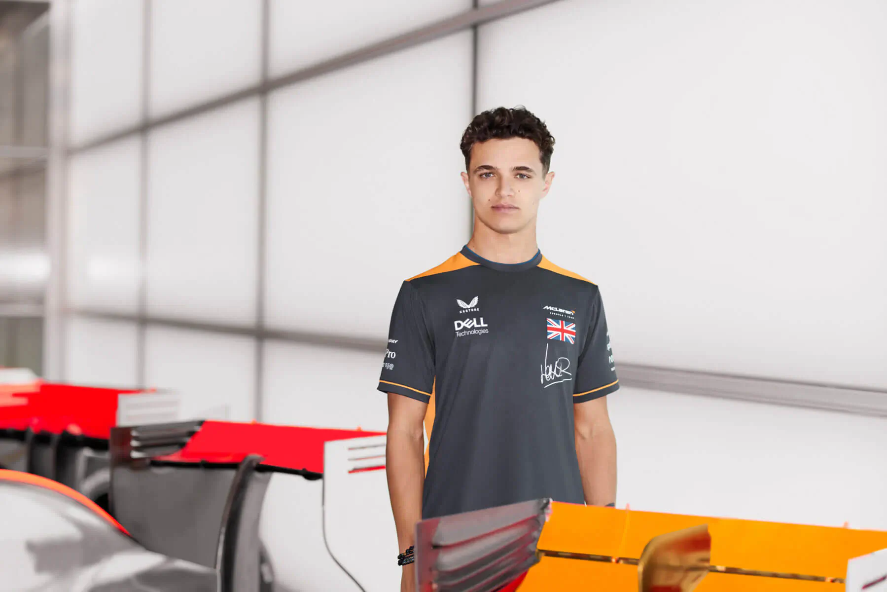 Young man in McLaren Castore black-orange polo stands by orange racing car in modern garage