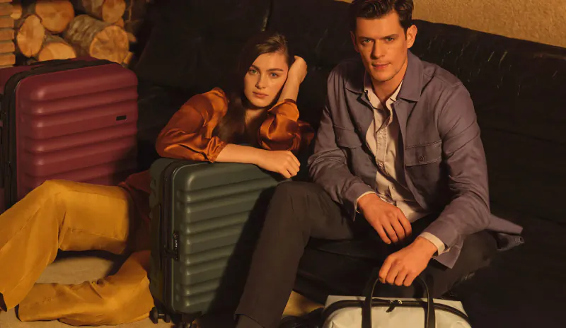 Smiling couple lounging on black sofa with Antler Clifton suitcases in maroon, green, and white duffel amid firewood.