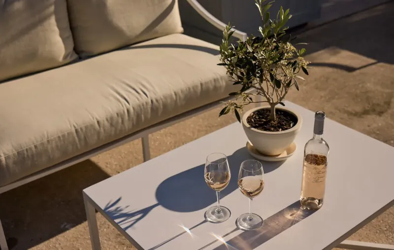 Two glasses of rosé wine and bottle on white table with olive tree pot, beige sofa, sunny terrace shadows.