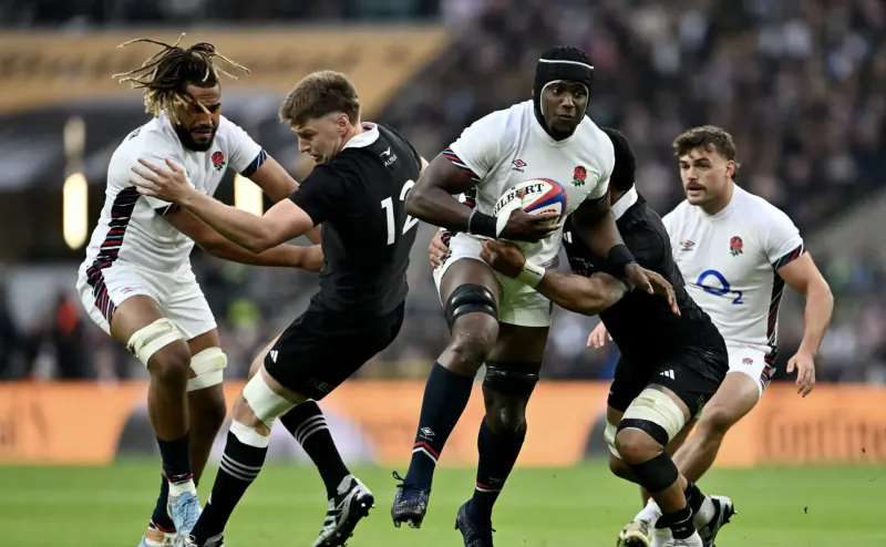 England rugby players in white tackle black-clad All Blacks player carrying ball on stadium field.