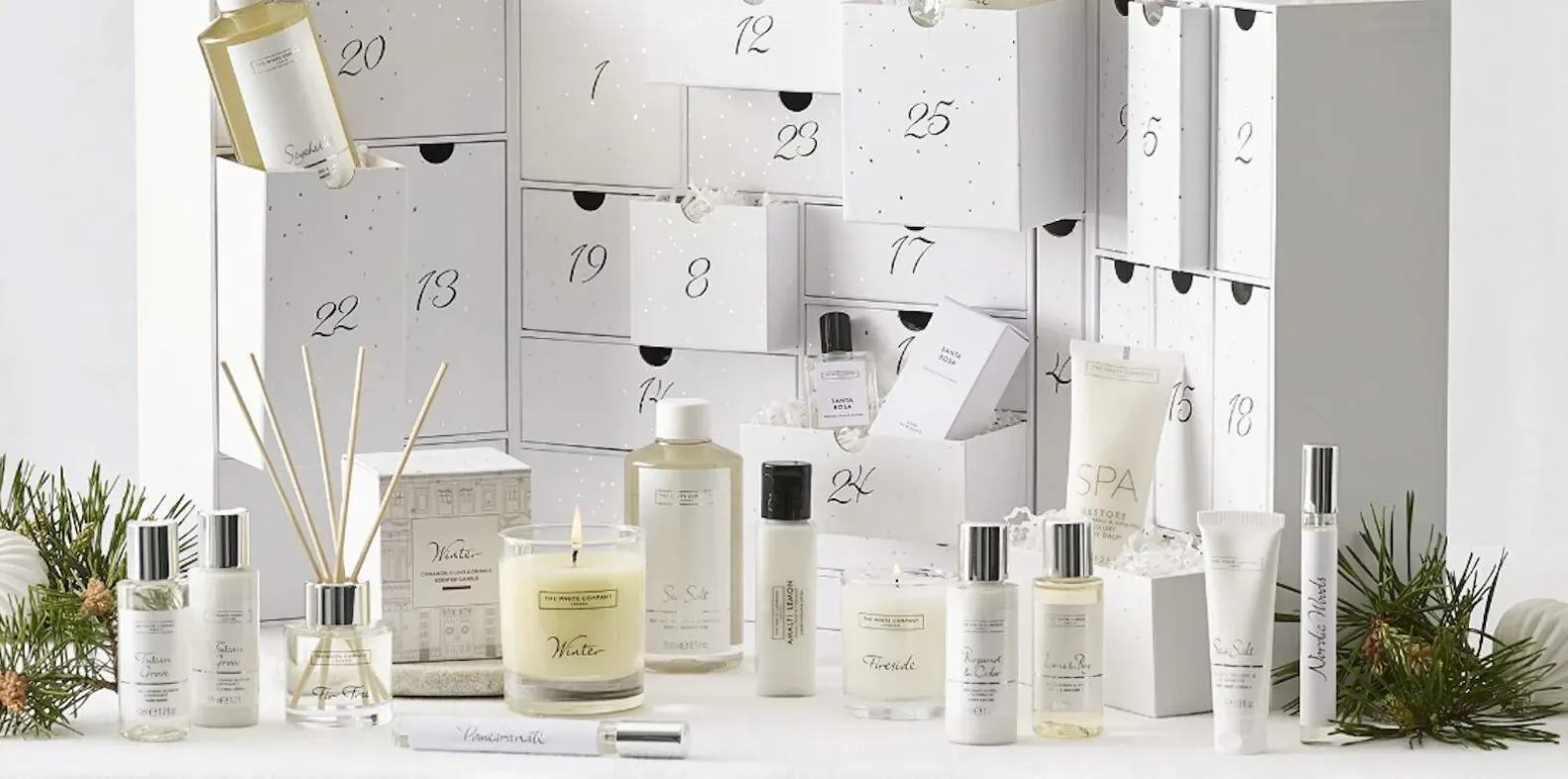 Luxury white advent calendar with numbered doors open, revealing skincare bottles, reed diffusers, candles, and fir branches.