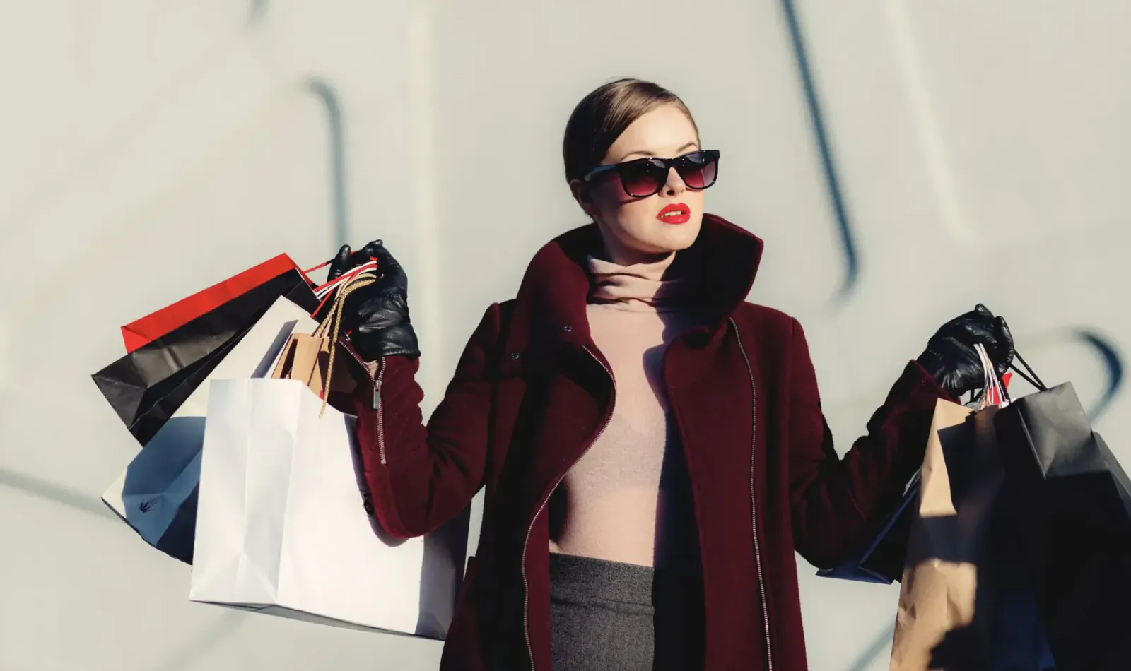 Stylish woman in maroon coat and sunglasses holding multiple shopping bags against gray wall
