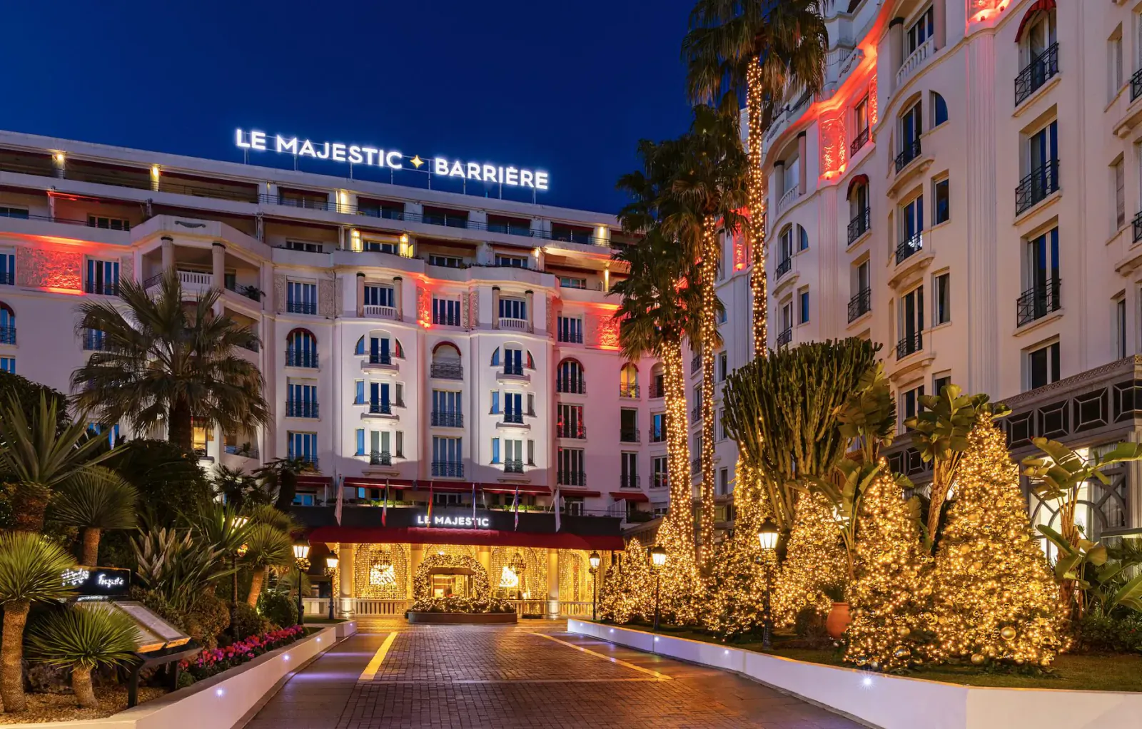 Le Majestic Barrière hotel in Cannes at night, lit up with red lights and Christmas trees at the entrance.