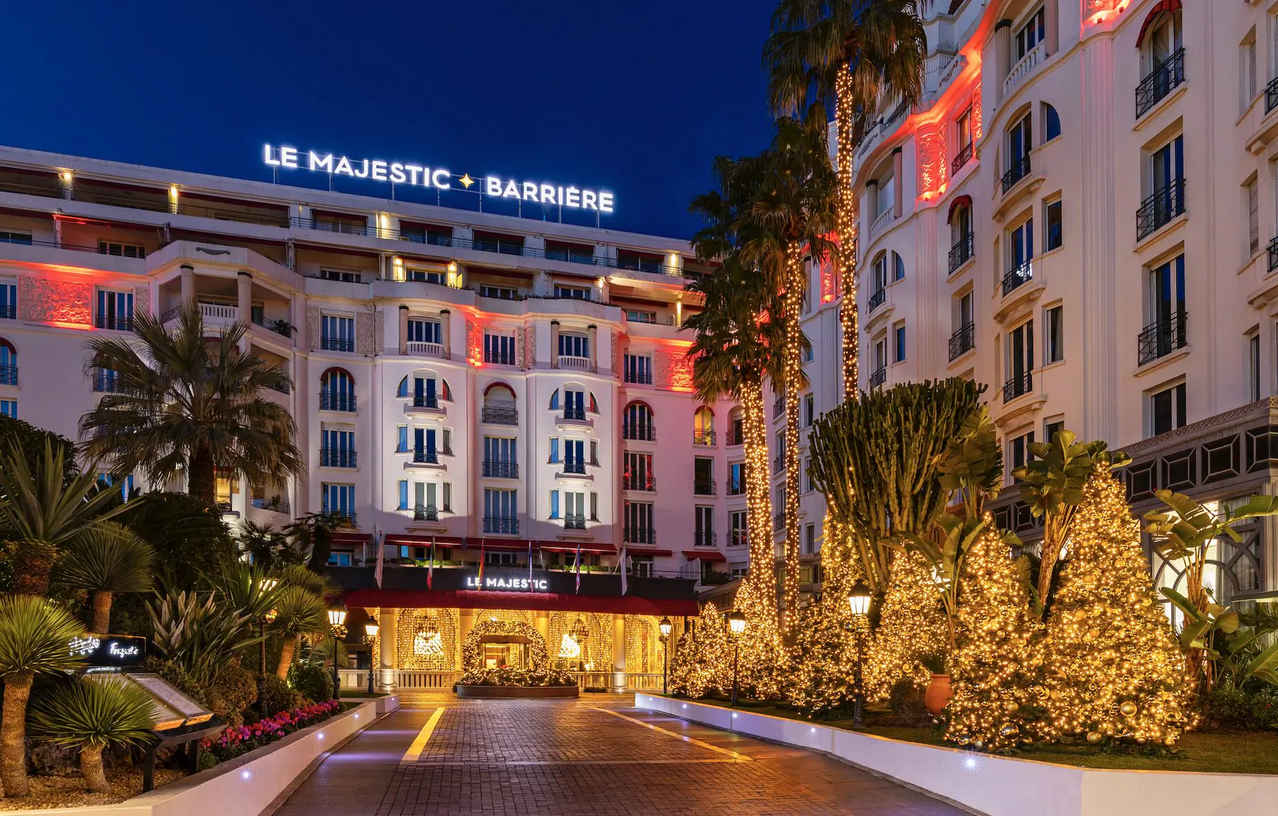 Le Majestic Barrière hotel in Cannes at night, lit up with red lights and Christmas trees at the entrance.