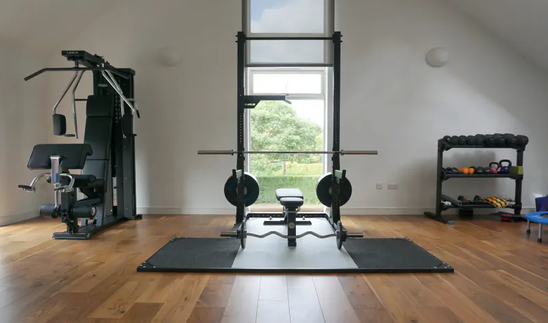 Home gym with Eleiko power rack, loaded barbell on bench, pull-up station, dumbbells, and large window.