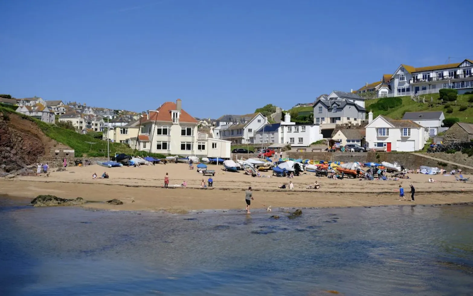 Sunny beach scene in South Devon with white hillside cottages, red-roofed building, cars, tents, and people by the sea.