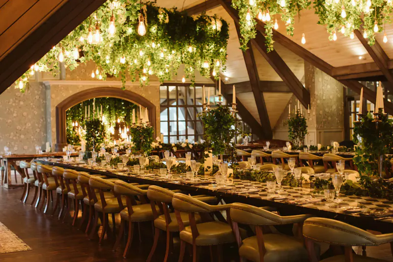Elegant wedding banquet table with greenery garlands, checkered linens, and hanging pendant lights in rustic beamed venue.
