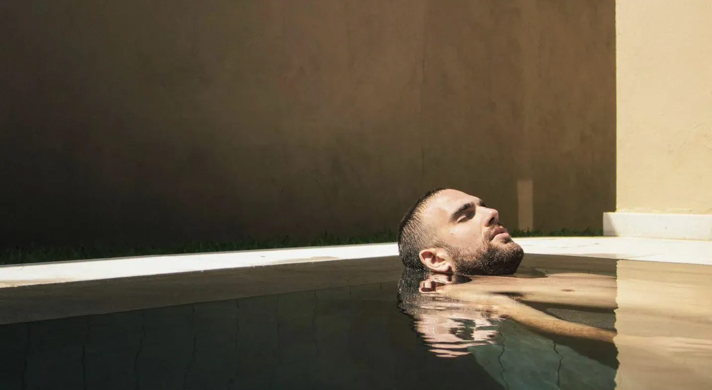 Bald man with beard reclining in luxurious indoor pool, head tilted back, warm lighting