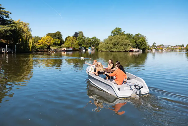 Group of four people, including a woman in orange, rowing a small motorboat on a calm river with trees and houses.