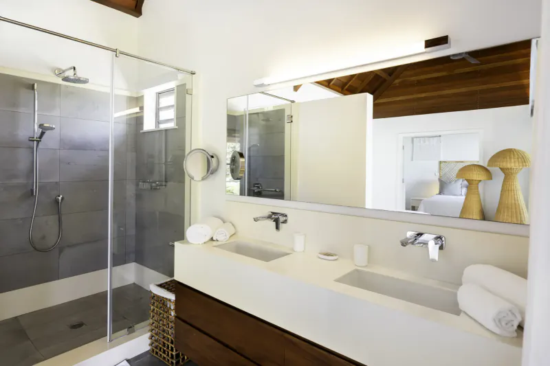 Modern bathroom in Villa Ruby, Mauritius villa: glass shower, double white sinks, mirror, woven lamps, wooden accents.