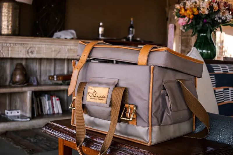 Gray Ahnasa safari cooler bag with leather straps on wooden table, beside wine bottles and flowers in rustic room