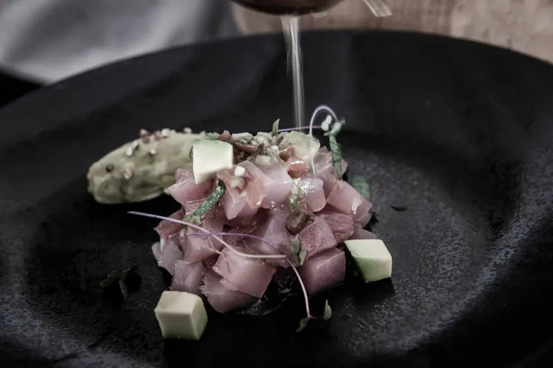Close-up of tuna tartare with avocado cubes, microgreens, and seeds on black plate, red wine pouring beside