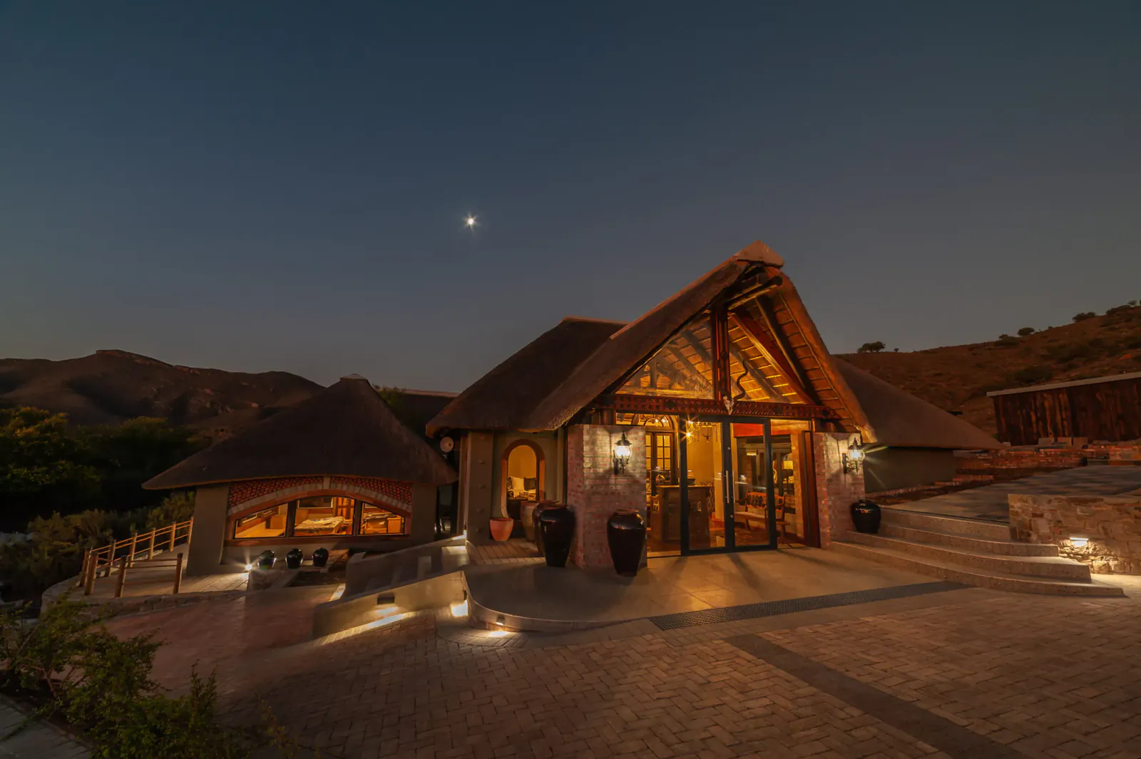 Twilight view of thatched Hippo arrival lodge at Mont Eco Game Reserve, lit warmly amid hills and starry sky.