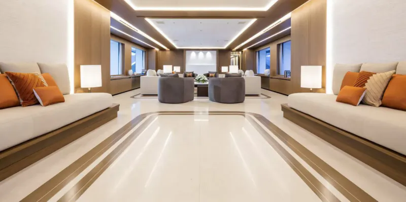 Modern luxury yacht lounge with white marble floors, orange cushions on low sofas, gray armchairs, and wood paneling.